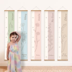 May include: Five vertical wall-mounted growth charts with wooden hangers, each featuring a unique floral line drawing, a name, and a measurement scale in feet. The charts display names Harper, Riley, Amelia, Mia Grace, and Liliana with flowers.