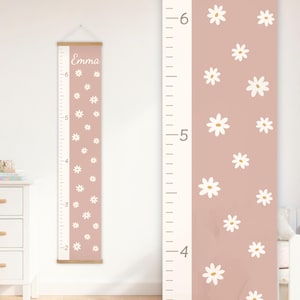 May include: A pink and white growth chart with daisy accents. The chart features a wooden top and bottom, with measurements in feet and inches. The name "Emma" is printed at the top. The chart is designed for tracking a child's height.