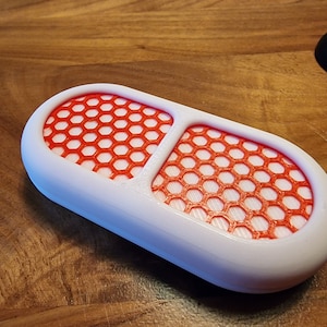 Op de afbeelding: Een witte, pilvormige container van plastic met twee compartimenten. Elk compartiment heeft een rood honingraatvormig gaas.