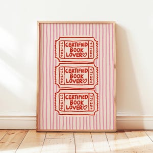 May include: A framed print with three red ticket stubs, each with the words "CERTIFIED BOOK LOVER" and a heart, on a pink and white striped background. The frame is light wood.