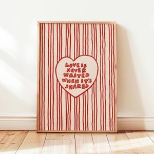 May include: A framed print with a cream background and vertical red stripes. A red heart in the center contains the text "LOVE IS NEVER WASTED WHEN IT'S SHARED" in red. The frame is a light brown color.