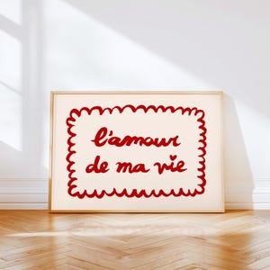 May include: A framed art print featuring the French phrase "l'amour de ma vie" in red script. The text is enclosed within a red scalloped border on a cream backdrop. The frame is a light wood colour.