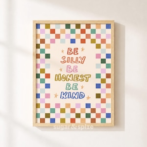 Op de afbeelding: Een rechthoekige wandkunstprint in een lichthouten lijst met een kleurrijke geblokte rand in tinten rood, oranje, geel, groen, blauw en roze. De tekst in het midden luidt BE SILLY BE HONEST BE KIND in veelkleurige letters met kleine sterretjes.