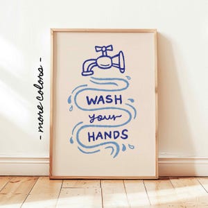 May include: A framed art print with a light beige background. The artwork features a blue illustration of a faucet with water droplets and the text "WASH your HANDS" in a whimsical font. The frame is a light wood color.