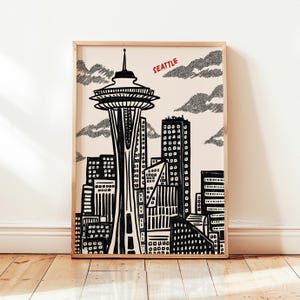 May include: Framed black and white art print of the Seattle skyline, featuring the Space Needle and other buildings. The word "SEATTLE" is printed in red. The artwork is displayed in a light-colored wooden frame.
