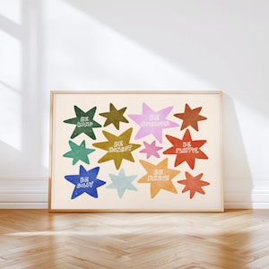 Op de afbeelding: Een ingelijste kunstprint met kleurrijke sterren met positieve affirmaties. De sterren zijn in verschillende kleuren, waaronder groen, blauw en oranje, en bevatten woorden als "Be Kind" of "Be Brave". De achtergrond is lichtbeige.