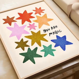 May include: A framed print featuring colorful star illustrations in shades of red, orange, pink, yellow, green, and blue. The words "YOU ARE MAGIC" are written in black. The artwork is displayed in a light wood frame.
