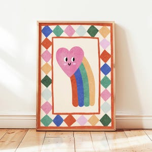 Può includere: Una stampa artistica incorniciata con un cuore rosa con un volto sorridente e un arcobaleno. L'opera d'arte è ambientata su uno sfondo color crema e incorniciata con un bordo di diamanti colorati e una cornice rossa. La stampa è esposta su un pavimento in legno.