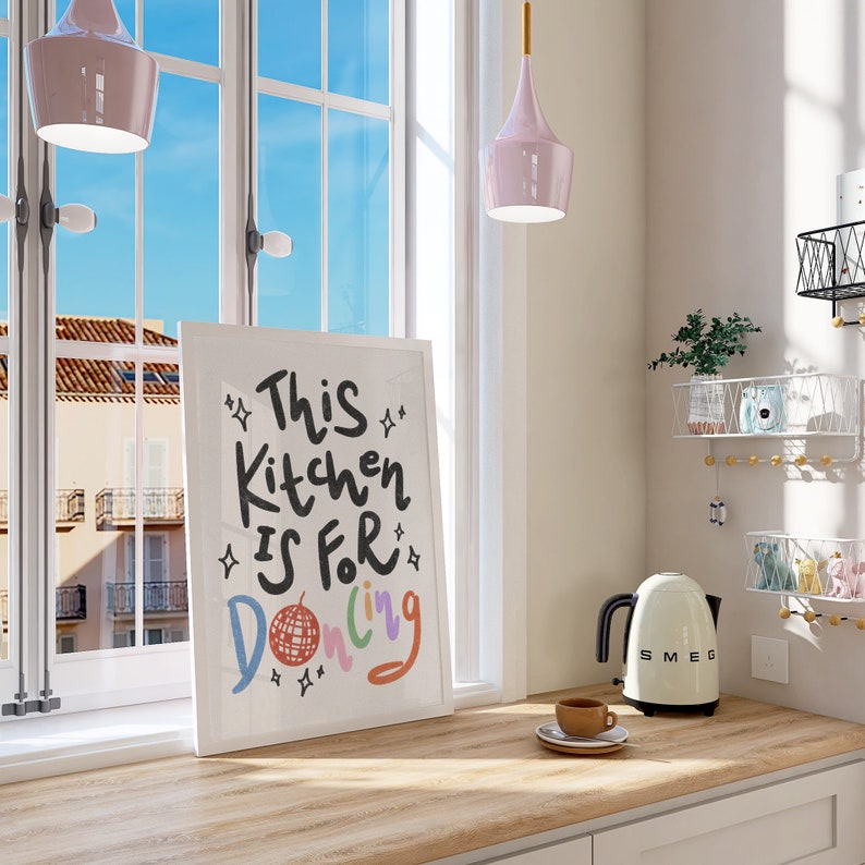 May include: A framed art print with the text "This Kitchen Is For Dancing" in a kitchen setting. The print is on a wooden countertop next to a white SMEG kettle and a cup of coffee. Two pink pendant lights hang from the ceiling.
