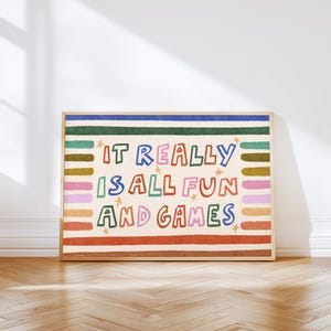 Peut inclure: Une affiche encadrée avec le texte "IT REALLY IS ALL FUN AND GAMES" dans des lettres colorées et ludiques. L'œuvre présente un fond crème avec des rayures horizontales et des étoiles, dans un cadre en bois.