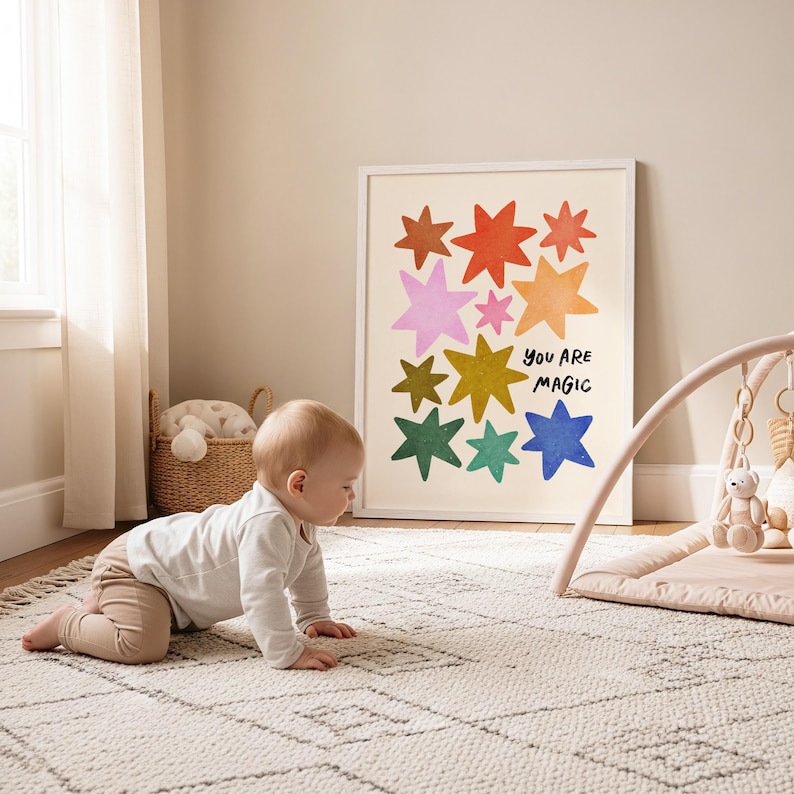 May include: A framed art print with colorful star illustrations and the words "You Are Magic" hangs on a wall. A baby crawls on a patterned rug in front of the artwork. A baby play gym and a woven basket are also in the room.