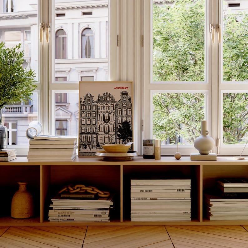 May include: A wooden shelf displays a framed Amsterdam print, stacks of books, a vase, and decorative items. The scene is set against a backdrop of large windows with a view of trees and buildings, creating a bright and airy atmosphere.