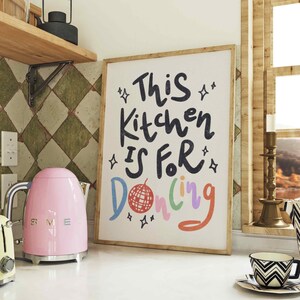 May include: A framed art print with the text "This Kitchen Is For Dancing" in black, with colorful accents. A pink electric kettle and a black and white patterned teacup are also visible.