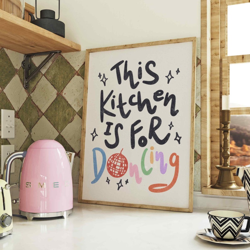 May include: A framed art print with the text "This Kitchen Is For Dancing" in black, with colorful accents. A pink electric kettle and a black and white patterned teacup are also visible.