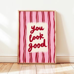 May include: A framed art print with a pink and red striped background. The print features the words "you look good" in red script on a cream-colored, scalloped-edged rectangle. The frame is a light wood color.
