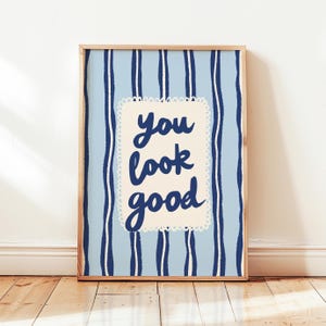 May include: A framed art print with a light blue striped background and the words "you look good" in dark blue script within a cream-colored, scalloped-edged rectangle. The frame is made of light-colored wood.
