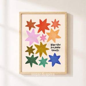 May include: A framed wall art print featuring a collection of multi-colored stars in shades of red, orange, yellow, green, blue, and pink on a cream background. The text "YOU CAN DO HARD THINGS" is printed in black, hand-lettered style font.