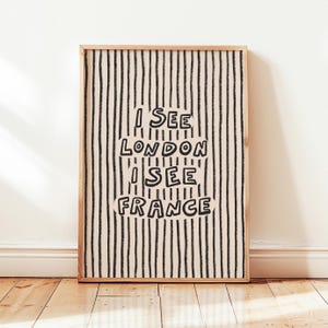May include: A framed art print with a light wood frame. The artwork features a cream-colored background with vertical black stripes. The text "I SEE LONDON I SEE FRANCE" is in a playful font.