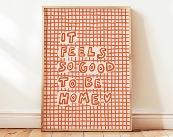 Könnte beinhalten: Ein gerahmter Kunstdruck mit dem Schriftzug "IT FEELS SO GOOD TO BE HOME" in orangefarbenen Buchstaben. Der Hintergrund ist ein Gittermuster in Orange und Weiß. Der Rahmen ist hellbraun. Das Kunstwerk wird an einer weißen Wand und einem Holzboden gezeigt.