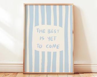 Puede incluir: Una impresión artística enmarcada con un marco de madera clara. La obra de arte presenta un fondo a rayas azul claro y crema con las palabras "THE BEST IS YET TO COME" en texto azul claro dentro de un óvalo crema. La impresión se muestra en un suelo de madera.