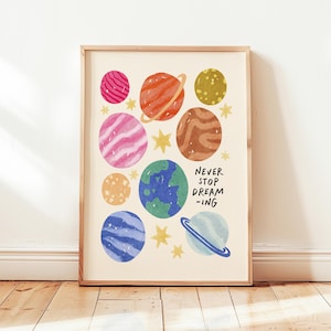 Puede incluir: Un estampado enmarcado con planetas y estrellas coloridas sobre un fondo crema. Los planetas son de color rosa, naranja, marrón, amarillo, azul y verde. El texto "NEVER STOP DREAM-ING" está impreso en negro.