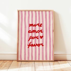 Peut inclure: Impression d'art encadrée avec un cadre en bois clair. L'œuvre présente des rayures verticales roses et blanches. Le texte rouge en cursive dit "more amor por favor" avec un symbole de cœur rouge. L'impression est affichée sur un sol en bois.