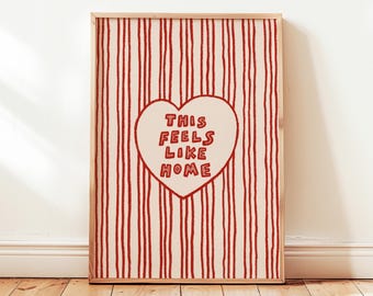 May include: A framed art print with a cream background and red vertical stripes. A cream-colored heart in the center contains the text "THIS FEELS LIKE HOME" in red. The frame is made of light-colored wood.