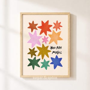 Puede incluir: Una lámina artística enmarcada con una colección de estrellas multicolores en tonos rojo, naranja, amarillo, rosa, verde y azul sobre un fondo crema. El texto "YOU ARE MAGIC" está impreso en negro, con una fuente manuscrita a la derecha.