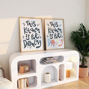 May include: Two framed art prints with gold frames, each displaying the text "This Kitchen Is For Dancing." One print features a black disco ball, while the other has a colorful disco ball. The prints are displayed on a white cabinet.