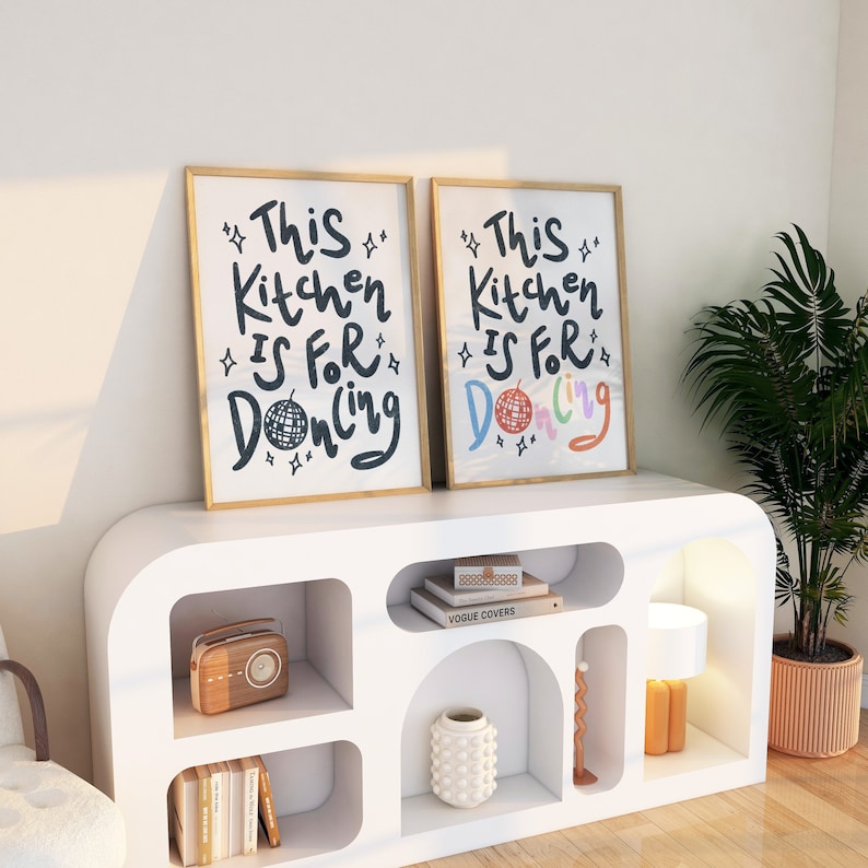 May include: Two framed art prints with gold frames, each displaying the text "This Kitchen Is For Dancing." One print features a black disco ball, while the other has a colorful disco ball. The prints are displayed on a white cabinet.