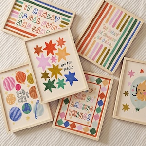 May include: A collection of framed art prints with colorful illustrations and inspirational quotes. The prints feature various designs, including stars, planets, and geometric patterns. Text includes "IT REALLY IS ALL FUN AND GAME", "YOU ARE MAGIC", and "CAN DO AMAZING THINGS".