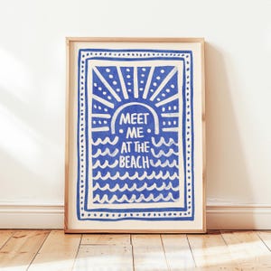 Puede incluir: Una impresión artística enmarcada con un diseño azul y blanco. La obra de arte presenta el texto "MEET ME AT THE BEACH" en una fuente estilizada, rodeado por un motivo de sol y olas. El marco es de color madera clara.