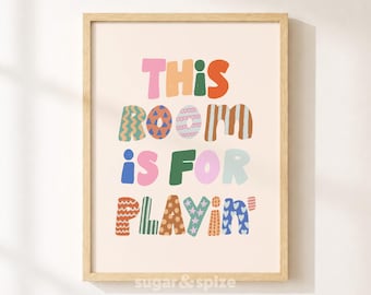 Op de afbeelding: Een ingelijste wandprint met de tekst THIS ROOM IS FOR PLAYIN' in kleurrijke, gedessineerde blokletters. De letters tonen diverse ontwerpen waaronder strepen, golven, stippen en sterren in tinten roze, oranje, groen en blauw.