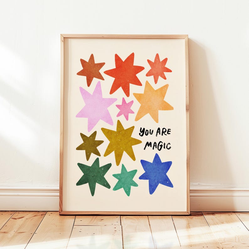 May include: A framed art print featuring colorful star illustrations in shades of red, orange, pink, yellow, green, and blue. The words "YOU ARE MAGIC" are written in black script. The artwork is displayed on a cream-colored background.