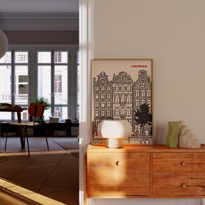 May include: A framed black and white print of Amsterdam buildings hangs above a wooden cabinet. A white mushroom-shaped lamp and two decorative vases sit on the cabinet. The room has hardwood floors and a view of a window.