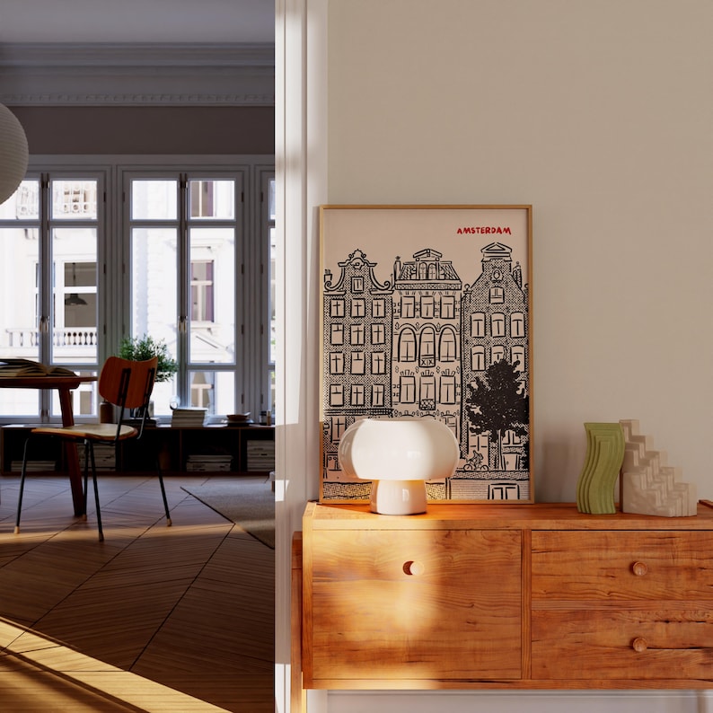 May include: A framed black and white print of Amsterdam buildings hangs above a wooden cabinet. A white mushroom-shaped lamp and two decorative vases sit on the cabinet. The room has hardwood floors and a view of a window.