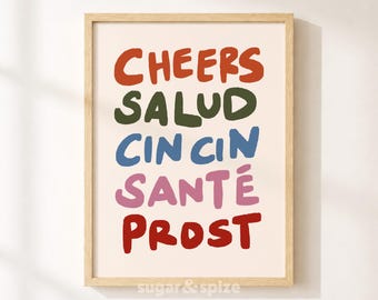 May include: A framed wall art print featuring the text CHEERS in orange, SALUD in dark green, CIN CIN in blue, SANTÉ in pink, and PROST in red, all set against a light cream background within a light-colored wooden frame.
