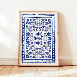 May include: A framed print featuring a blue floral pattern and the text "HOME SWEET HOME". The artwork is set against a cream background and framed in light wood. The print is displayed on a wooden floor.