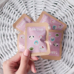 Op de afbeelding: Een set van vijf kleine bruine zakjes met roze en witte illustraties van Sanrio-karakters. De zakjes zijn gelabeld met "Sanrio" en bevatten karakters zoals Hello Kitty, Keroppi en My Melody.