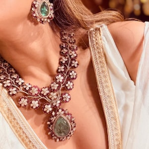 May include: Elegant jewelry set featuring a necklace and earrings. The necklace and earrings have a floral design with white and red accents, and a large green gemstone pendant. The earrings have a teardrop shape.