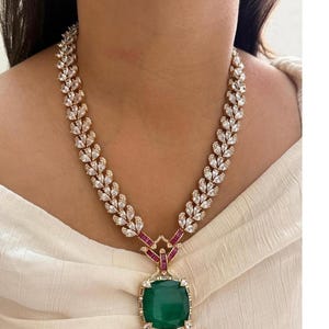 May include: A gold-toned necklace with a leaf-like design, adorned with clear crystals. The necklace features a large, square, emerald-green pendant with a surrounding of small crystals and red accents.