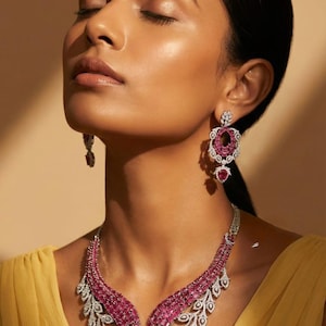 May include: A stunning jewelry set featuring a necklace and earrings. The necklace has a central pendant and a cascading design with pink gemstones and white accents. The earrings mirror the necklace design. The jewelry is set against a yellow dress.