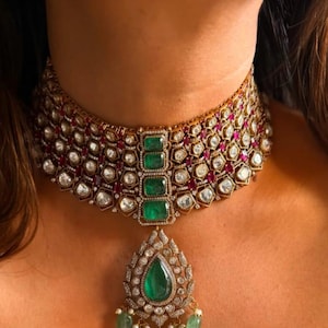 Może przedstawiać: Złoty naszyjnik z akcentami szmaragdów i rubinów. Naszyjnik typu choker posiada centralną zawieszkę z dużą szmaragdową łzą i mniejszymi szmaragdowymi kroplami. Luksusowa biżuteria.