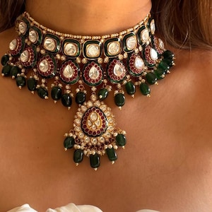 Może przedstawiać: Złoty naszyjnik typu choker z szmaragdowo-zielonymi i rubinowo-czerwonymi akcentami. Naszyjnik posiada kwadratowe i łezkowate kamienie szlachetne, z centralną zawieszką. Projekt jest bogaty i szczegółowy, o klasycznej estetyce.