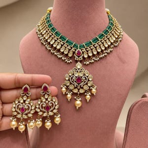May include: A necklace and earring set featuring multiple strands of green beads, gold-tone metal accents, clear stones, and teardrop-shaped pink center stones. The necklace includes a large central pendant, while the earrings match the design.