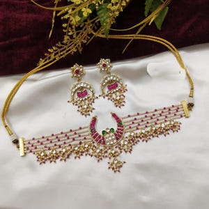 May include: An ornate jewelry set featuring a choker necklace and matching earrings. The necklace and earrings have a gold-tone metal base, adorned with pearls, red and green gemstones, and a crescent-shaped pendant with a floral design.
