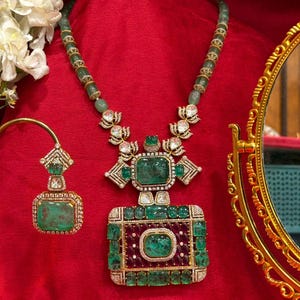 May include: An opulent emerald and diamond jewellery set, comprising a necklace and earrings, presented on a red backdrop. The necklace showcases a substantial rectangular pendant with a central emerald, encircled by smaller emeralds and diamonds. The earrings echo this design.