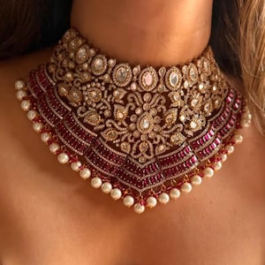 Peut inclure: Un collier ras du cou doré orné de motifs floraux et géométriques complexes. Le collier est orné de pierres précieuses transparentes et rouge rubis, bordé de petites perles blanches. Le design est symétrique et orné, idéal pour les occasions formelles.
