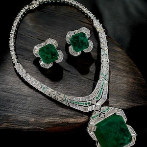 Può includere: Un lussuoso set di gioielli con collana e orecchini abbinati. La collana e gli orecchini sono adornati con grandi smeraldi a taglio quadrato e circondati da un'incastonatura scintillante simile a diamanti. I gioielli sono esposti su una superficie di legno scuro.