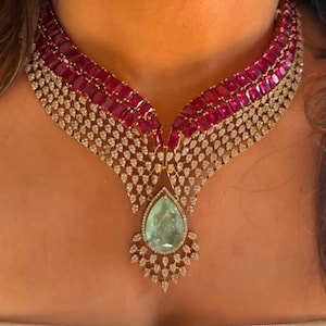 May include: An opulent necklace with a gold setting, adorned with rows of red gemstones and diamonds. The necklace features a large, pear-shaped, light green gemstone pendant. The design is intricate and elegant, suitable for special events.